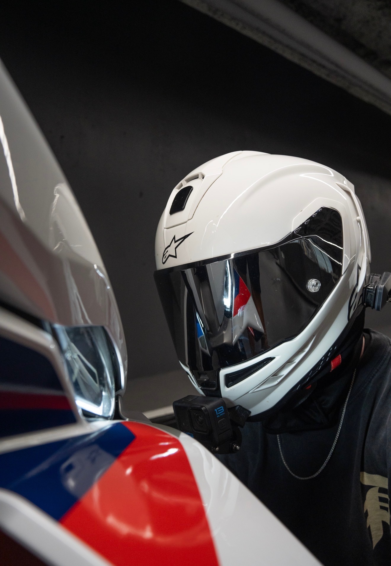 Close-up of Blaze's white full-face helmet with mirrored silver visor, reflecting the BMW M colors of his motorcycle, in a dark parking garage.