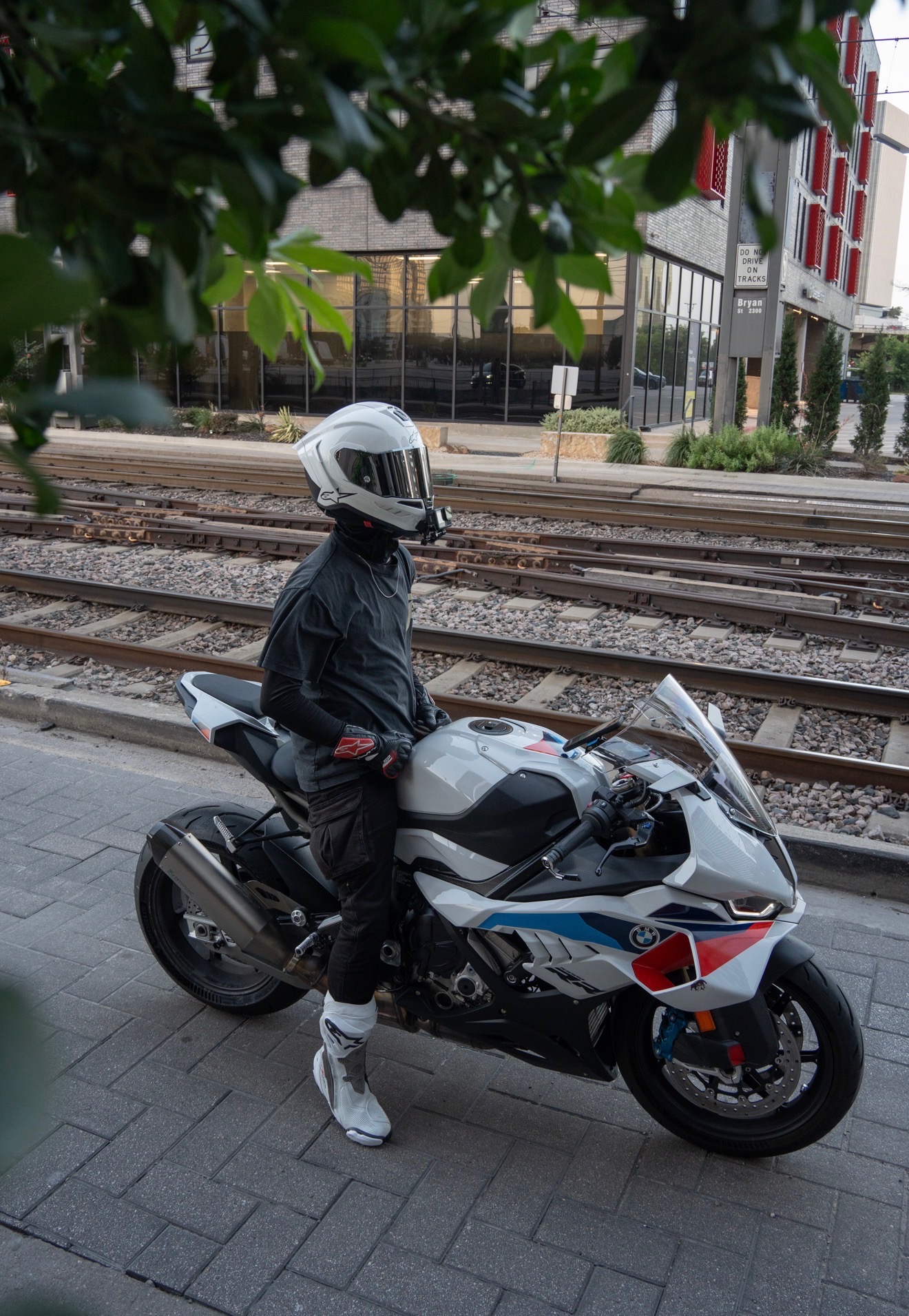 Blaze standing beside his white, blue, and red BMW S1000RR near light rail tracks in downtown Dallas at dusk.