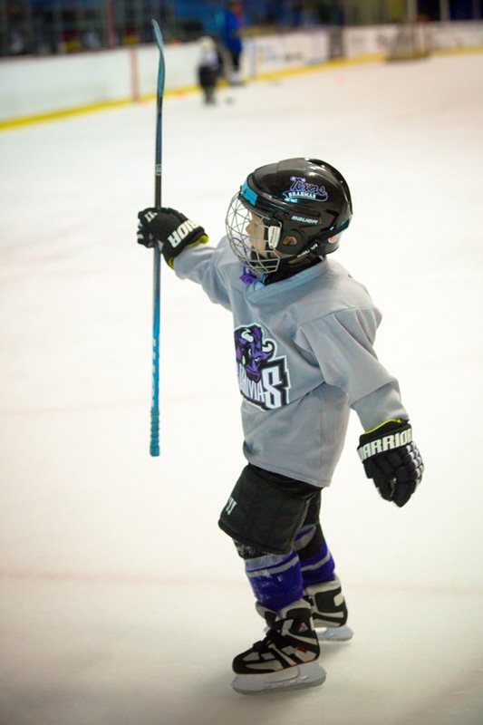 A young hockey player in full cage-helmet gear celebrating on the ice, stick raised, at NYTEX rink.