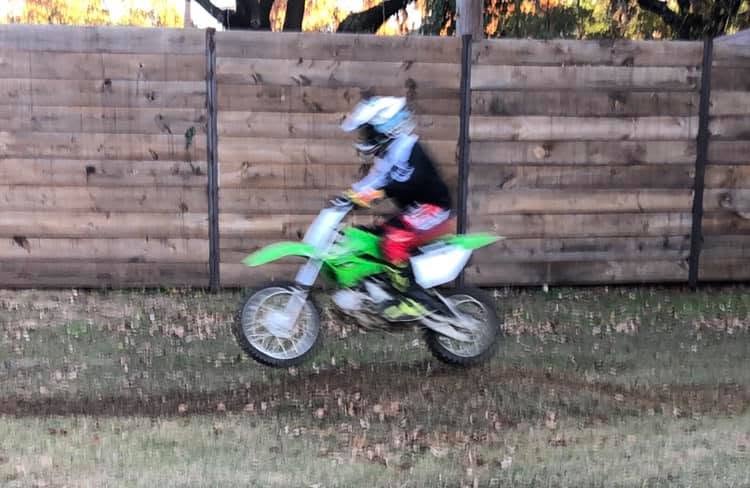 Blaze catching air on a green Kawasaki dirt bike along a wooden fence line, motion-blurred.