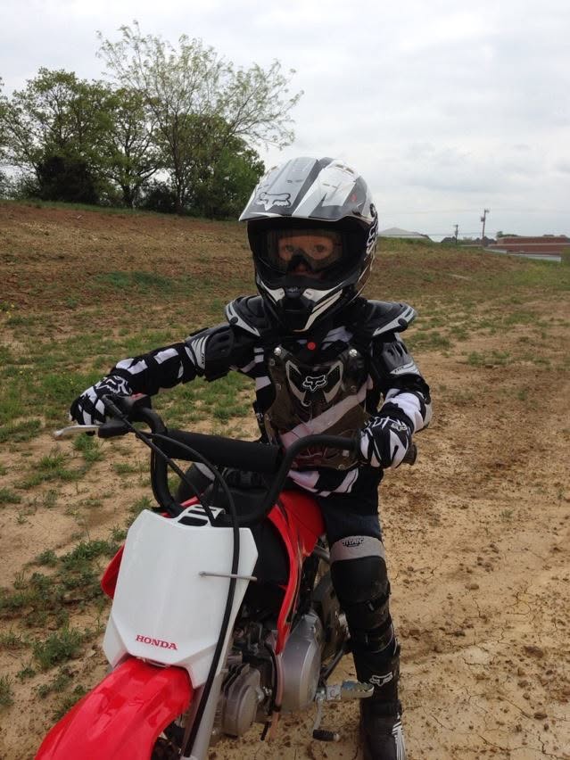 Young Blaze on a red Honda 50 dirt bike wearing full motocross gear, standing in a Texas field.