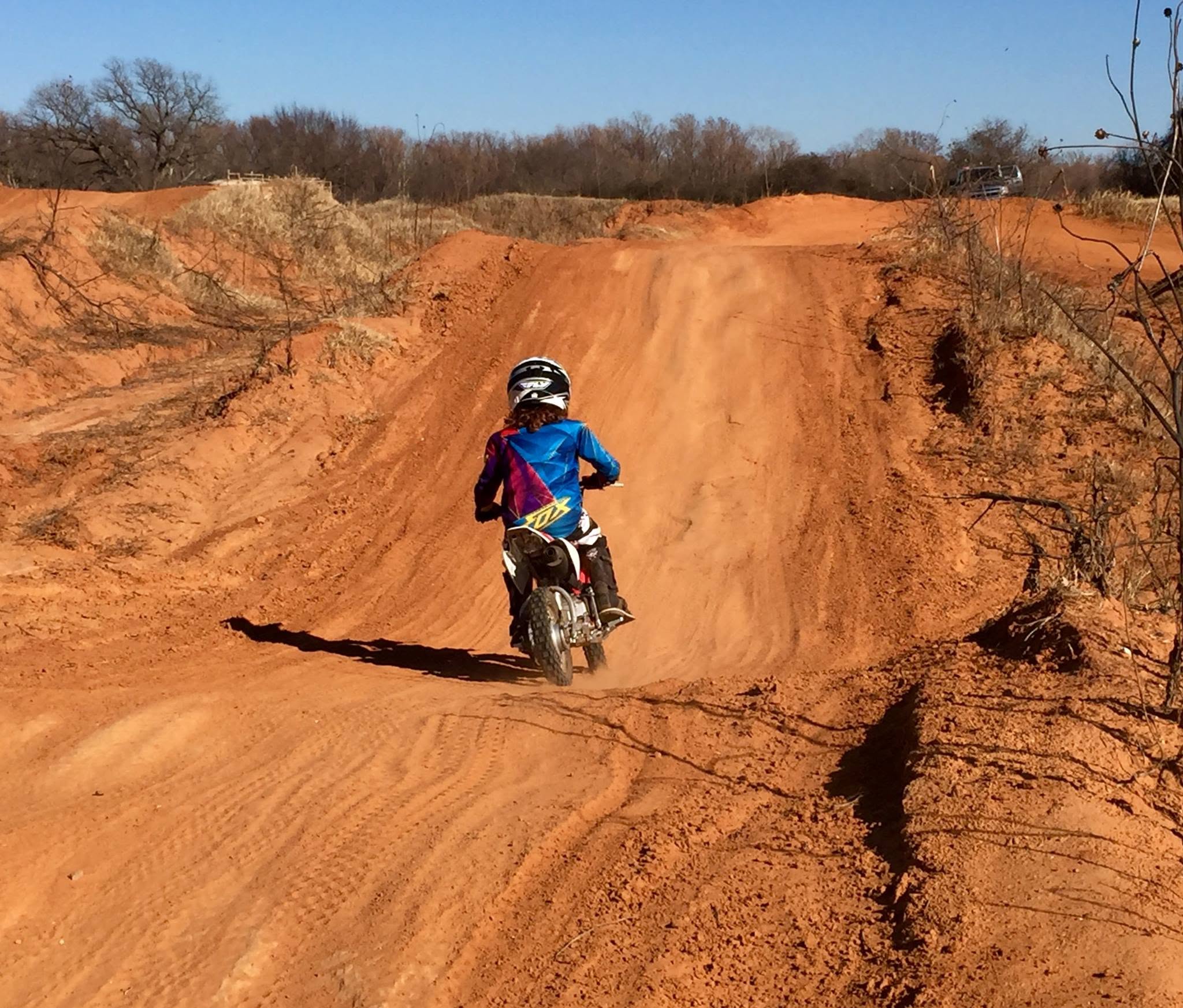 Blaze riding a small motocross bike over a dirt berm on a red-clay motocross track.
