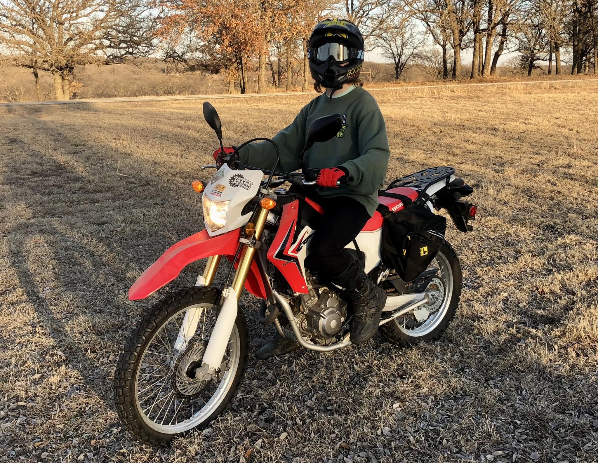 Blaze sitting on a red and white Honda CRF250L dual-sport motorcycle in a dry winter field, packed for a ride.