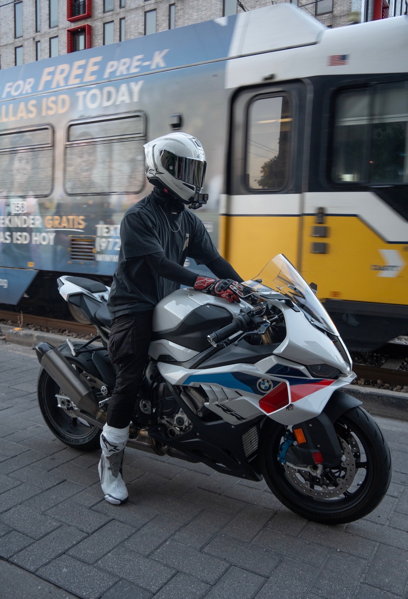 Blaze, helmet on, standing beside his white and BMW M-colored S1000RR as a DART train passes behind him on the tracks in downtown Dallas.
